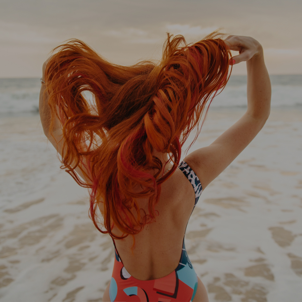 young red-haired woman with flying hair on the ocean coast at sunset, rear view
